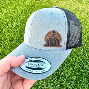 May include: A gray snapback hat with a black mesh back and a brown leather patch featuring a circus tent design. The hat has a curved brim and a white tag that reads "Snapback" and "Yupoong Since 1974".