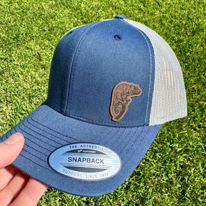May include: A navy blue Snapback hat with a white mesh back and a brown leather patch featuring a chameleon design. The hat has a curved brim and the text "THE AUTHENTIC YUPOONG SNAPBACK" on the front.