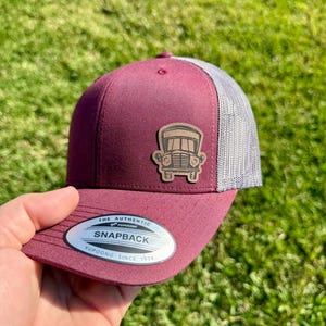 May include: Maroon and gray snapback hat with a wooden patch featuring a bus design. The hat has a mesh back and a curved brim. The hat has a tag that reads "The Authentic Snapback Yupoong Since 1974".