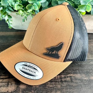 May include: A tan Snapback baseball cap with a black mesh back and a black wolf silhouette patch. The cap has a curved brim and a white and black tag that reads "The Authentic Snapback Yupoong Since 1974."