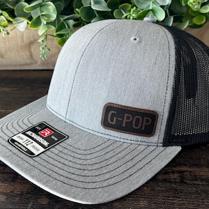 May include: A gray Richardson 112 trucker hat with a black mesh back. The hat features a brown leather patch with the text "G-POP" in brown letters. The brim is gray with black stitching.