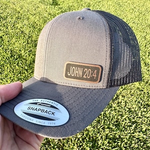May include: Gray trucker hat with a black mesh back and a curved brim. The hat features a leather patch with the text "JOHN 20:4". The hat also has a white and black sticker that says "SNAPBACK".
