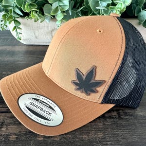 May include: A brown and black snapback hat with a cannabis leaf patch. The hat features a mesh back and a curved brim. The front of the hat has the text "THE AUTHENTIC SNAPBACK YUPOONG SINCE 1974".