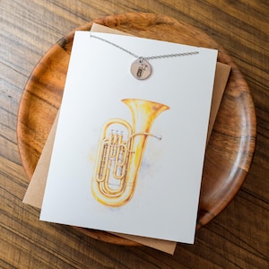 May include: A watercolor illustration of a golden tuba on a white card, resting on a wooden plate. A silver necklace with a small, round pendant featuring a tuba design hangs above the card. The background is a wooden surface.