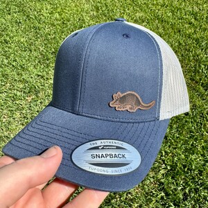 May include: Navy blue snapback hat with a white mesh back and a brown armadillo patch. The hat features the text "THE AUTHENTIC YUPOONG SNAPBACK YUPOONG SINCE 1974" on a silver oval patch.