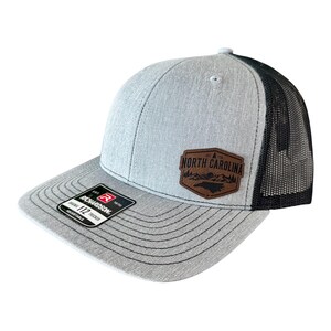 May include: A gray trucker hat with a black mesh back. The hat features a brown leather patch with the words "North Carolina" and a mountain and tree design. The brim is gray with black stitching, and the hat is adjustable.