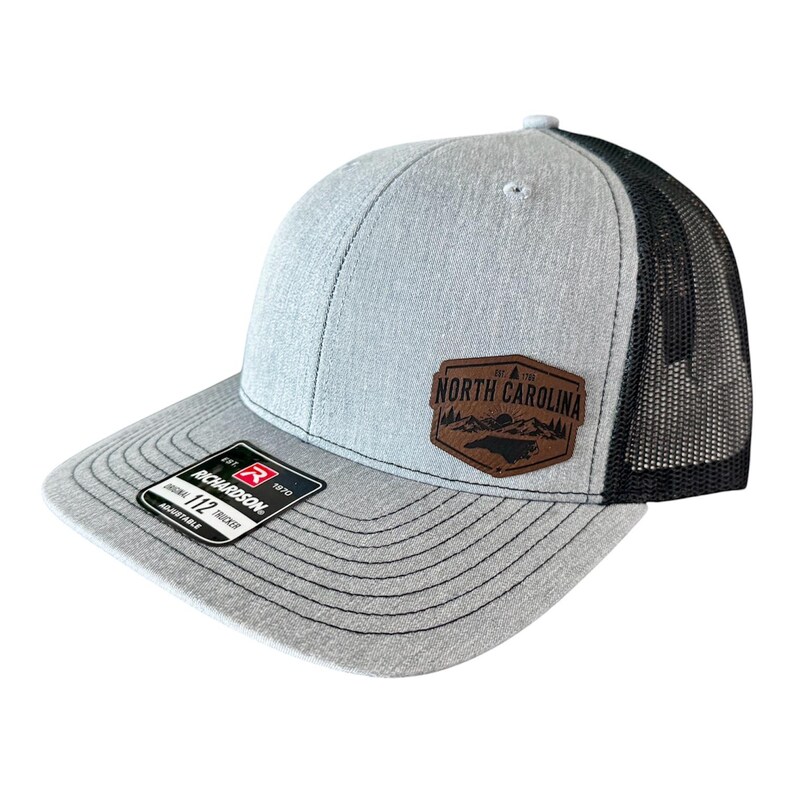 May include: A gray trucker hat with a black mesh back. The hat features a brown leather patch with the words "North Carolina" and a mountain and tree design. The brim is gray with black stitching, and the hat is adjustable.