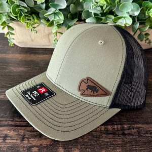 May include: An olive green baseball cap with a black mesh back, featuring a brown leather-like patch shaped like an arrowhead with a black bison silhouette. A black sticker on the brim displays the text 112 The Original MD-LG.