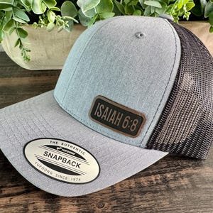 May include: A gray Snapback baseball cap with a black mesh back. The cap features a leather patch with the text "ISAIAH 6:8". The hat has a curved brim and a Yupoong tag.