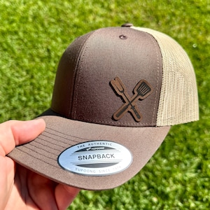 May include: Brown and tan trucker hat with a leather patch featuring crossed grilling utensils. The hat has a curved brim and a mesh back. The patch is brown with black detailing. The hat has a snapback closure and a silver and black tag that reads "Snapback".