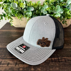 May include: A heather gray trucker hat with a black mesh back and a flat brim. The front features a brown leather patch embossed with an all-terrain vehicle graphic. A black sticker on the brim displays the text Richardson 112 Adjustable.
