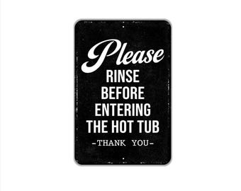 Peut inclure: Un panneau rectangulaire en métal noir aux coins arrondis avec le texte blanc : Please RINSE BEFORE ENTERING THE HOT TUB -THANK YOU-. Le panneau présente une texture légèrement vieillie et deux petits trous de fixation en haut et en bas.