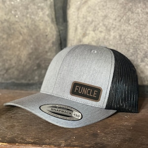 May include: A gray and black trucker hat with the word "FUNCLE" on a brown patch. The hat has a curved brim and a mesh back. The hat also has a "Yupoong Snapback" tag.