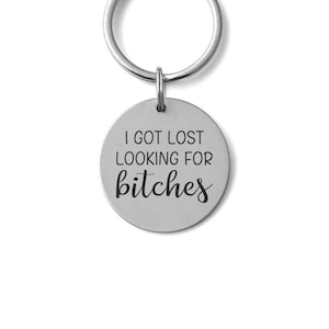 May include: A silver-colored keychain with a round pendant. The pendant has the phrase "I GOT LOST LOOKING FOR bitches" in black, stylized text. The keychain ring is also silver.
