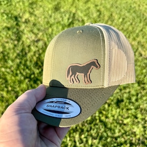 May include: Olive green and tan trucker hat with a horse silhouette patch. The hat features a mesh back, a curved brim, and a snapback closure. The tag reads "The Authentic Snapback Yupoong Since 1974".
