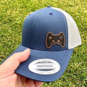 May include: Navy blue snapback hat with a white mesh back and a brown leather patch featuring a video game controller. The hat has a curved brim and the text "THE AUTHENTIC SNAPBACK" on the front.