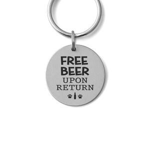 May include: A silver-colored keychain tag with the text "FREE BEER UPON RETURN" in bold, black letters. Below the text are paw prints and a beer bottle graphic. The tag is attached to a silver key ring.