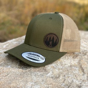 May include: Olive green and tan trucker hat with a curved brim. The hat features a round, brown leather patch with a forest and Bigfoot silhouette. The tag reads "THE AUTHENTIC SNAPBACK YUPOONG SINCE 1974".