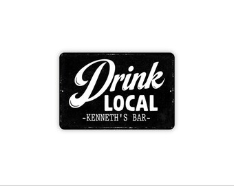 May include: A rectangular black metal sign with rounded corners featuring the white text Drink Local in a stylized script and block font. Below this, the text -KENNETH'S BAR- is printed in a smaller white block font. The sign has a distressed finish.