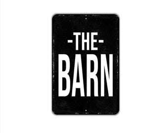 May include: A rectangular black metal sign with rounded corners and a distressed texture. The sign features the white text -THE- centered above the word BARN in a large, bold, sans-serif font. There are two small mounting holes visible.
