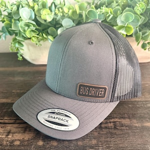 May include: Gray trucker hat with a black mesh back and a curved brim. The hat features a brown leather patch that reads "BUS DRIVER". The hat also has a white and black tag that reads "THE AUTHENTIC SNAPBACK YUPOONG SINCE 1974".