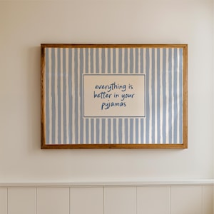 May include: A framed print with a light blue and white striped background. The print features a rectangular inset with the text "everything is better in your pyjamas" in a blue font. The frame is a warm brown colour.