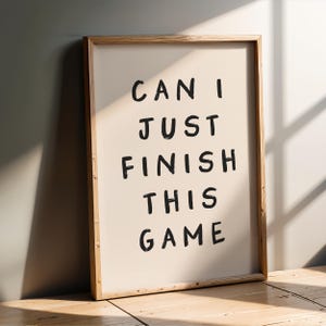 May include: A framed print with the text "CAN I JUST FINISH THIS GAME" in black handwritten font on a cream background.