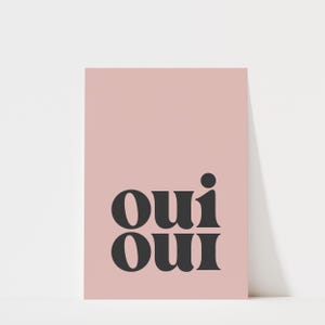 May include: A rectangular wall art print featuring a solid dusty pink background with the word "oui" printed twice in a stacked format using a bold, dark charcoal gray serif typeface with rounded, stylized letterforms.