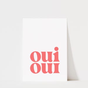 May include: A rectangular white print featuring the word "oui" repeated twice in a stacked format. The text is rendered in a bold, coral-pink serif typeface, with the two instances of the word aligned vertically in the center of the white background.