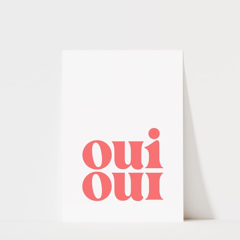 May include: A rectangular white print featuring the word "oui" repeated twice in a stacked format. The text is rendered in a bold, coral-pink serif typeface, with the two instances of the word aligned vertically in the center of the white background.