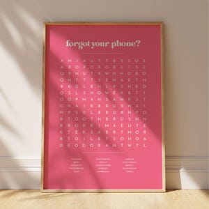 May include: A framed pink word search puzzle with the phrase "forgot your phone?" in white text at the top. The puzzle contains words related to bathroom items. A wooden frame surrounds the print.