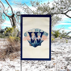 May include: A garden flag with a navy blue border and a seashell design. The seashell is blue and white with the word "Williams" in a script font. The flag is displayed outdoors against a backdrop of trees and a blue sky.