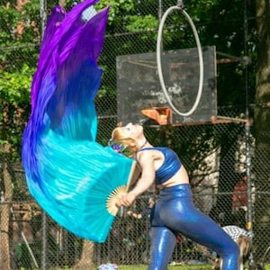 May include: A woman wearing a blue and purple silk scarf, a blue crop top, and blue sparkly leggings is performing with a fan and a hoop. She is standing in front of a basketball hoop and a chain link fence.