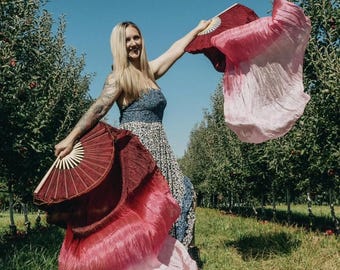 May include: A woman in a floral dress holds two silk fans in an orchard. One fan is burgundy and the other is pink and white. The fans are open, with the silk flowing in the wind. The background is a sunny day with rows of trees.