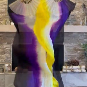 May include: A pair of open fans with a flowing fabric in the colors of purple, yellow, white, and black. The fans are held open, displaying the vibrant colors of the fabric. The fabric appears to be silk or a similar lightweight material.