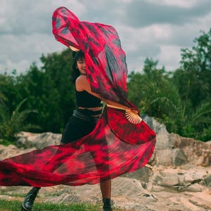 May include: A woman wearing a black outfit and holding a red and black fabric, which is flowing in the wind. She is standing in a rocky outdoor setting with green trees in the background.