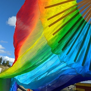May include: A colourful rainbow silk fan against a clear blue sky. The fan displays a smooth colour gradient from red to orange, yellow, green, blue, and purple. Wooden ribs support the fabric, creating a fanned-out shape.