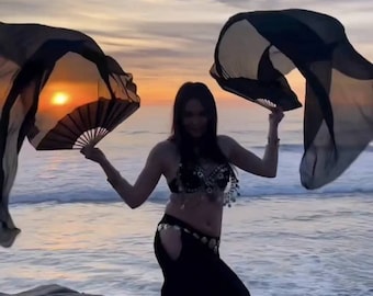 May include: A woman wearing a black costume with a silver belt dances on a beach at sunset. She is holding two black fans and is twirling them in the air.
