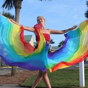 May include: A person is holding a large, flowing rainbow-colored silk fan. The fan's colors transition smoothly from red to purple. The person is wearing a red top and is standing outside on a sunny day.