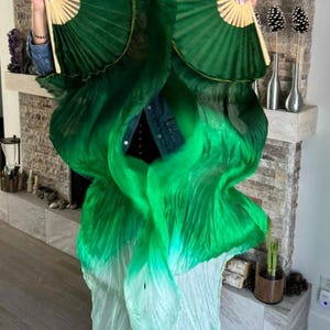 May include: A green and white silk dance veil with a gradient effect. The veil is being held by a person, who is also holding two green silk fans.