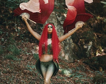 May include: A person in a forest setting, wearing a costume with red hair and holding two red and white fans. The costume includes a green top and bottom with leaf-like details. The fans have a dark red frame and a light red and white fabric.