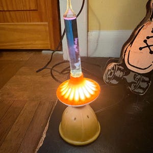 May include: A vintage mushroom-shaped lamp with a clear, cylindrical glass tube filled with liquid. The lamp has an orange, flower-like shade and a yellow base. The lamp is plugged in and illuminated, casting a warm glow.