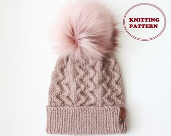 May include: A pink knitted beanie with a large fluffy pom pom on top. The beanie has a cable knit pattern and a leather tag with the word "Thread &" on it.