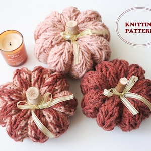 May include: Three knitted pumpkins in shades of pink and brown with a beige ribbon tied around each stem. The pumpkins are on a white surface.  KNITTING PATTERN is written in red on a white circle.