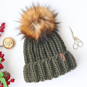 May include: A green knitted beanie with a large brown and black faux fur pom pom. The beanie has a brown leather tag with the word "Knit" on it.