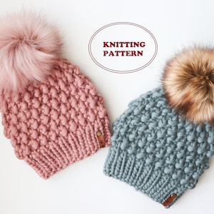 Puede incluir: Dos gorros de punto con pompones de piel sintética. Un gorro es de color rosa polvoriento y el otro es de color azul claro. Los gorros están hechos con un hilo grueso y tienen un patrón de punto texturizado. El texto "Knitting Pattern" es visible en la imagen.