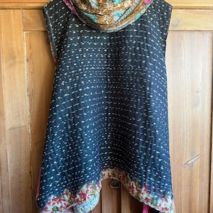 May include: A sleeveless tunic featuring a dark navy blue quilted fabric with small white horizontal dashes. The hem displays a colourful floral pattern in pink, green, and white. A matching scarf with a brown, teal, and pink paisley print is draped around the neck.