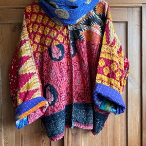 May include: A quilted jacket featuring a vibrant patchwork of red, yellow, black, and blue fabric panels with intricate kantha stitching. The garment has wide sleeves with rolled cuffs and a matching blue patterned scarf draped around the collar.