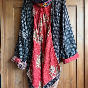 May include: A long-sleeved quilted jacket featuring a dark exterior with a repeating black and white paisley pattern and a contrasting interior lining in bright red with a floral rose print. A matching scarf is draped over the top collar.