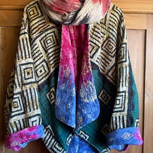 May include: A quilted jacket featuring a geometric pattern of black and cream squares on the outer panels, with a vibrant pink, blue, and green floral interior lining. The garment includes a matching scarf draped around the collar area.